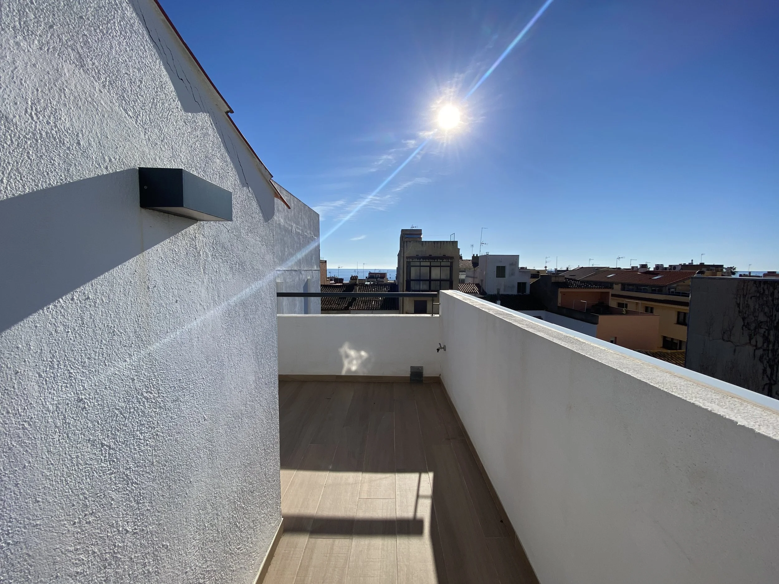 Vista de una terraza con suelo de madera, paredes blancas y un muro con una barandilla, con el sol brillante en un cielo despejado y edificios residenciales al fondo.
