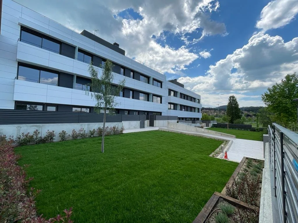 Edificio moderno de varias plantas con ventanas grandes y fachada blanca, con área verde con césped bien cuidado, un árbol joven en el centro, y caminos pavimentados, bajo un cielo con nubes y sol.