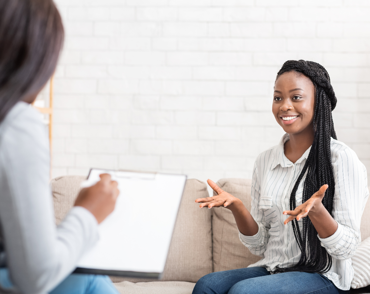 Une jeune femme souriante assise sur un canapé, échangeant de manière dynamique avec une psychologue du travail tenant un presse-papiers. La scène illustre un accompagnement personnalisé et bienveillant pour l'orientation professionnelle des jeunes