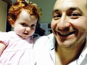 Margot and Margot's Dad smiling for a selfie indoors, with the girl wearing a pink dress and having curly red hair.