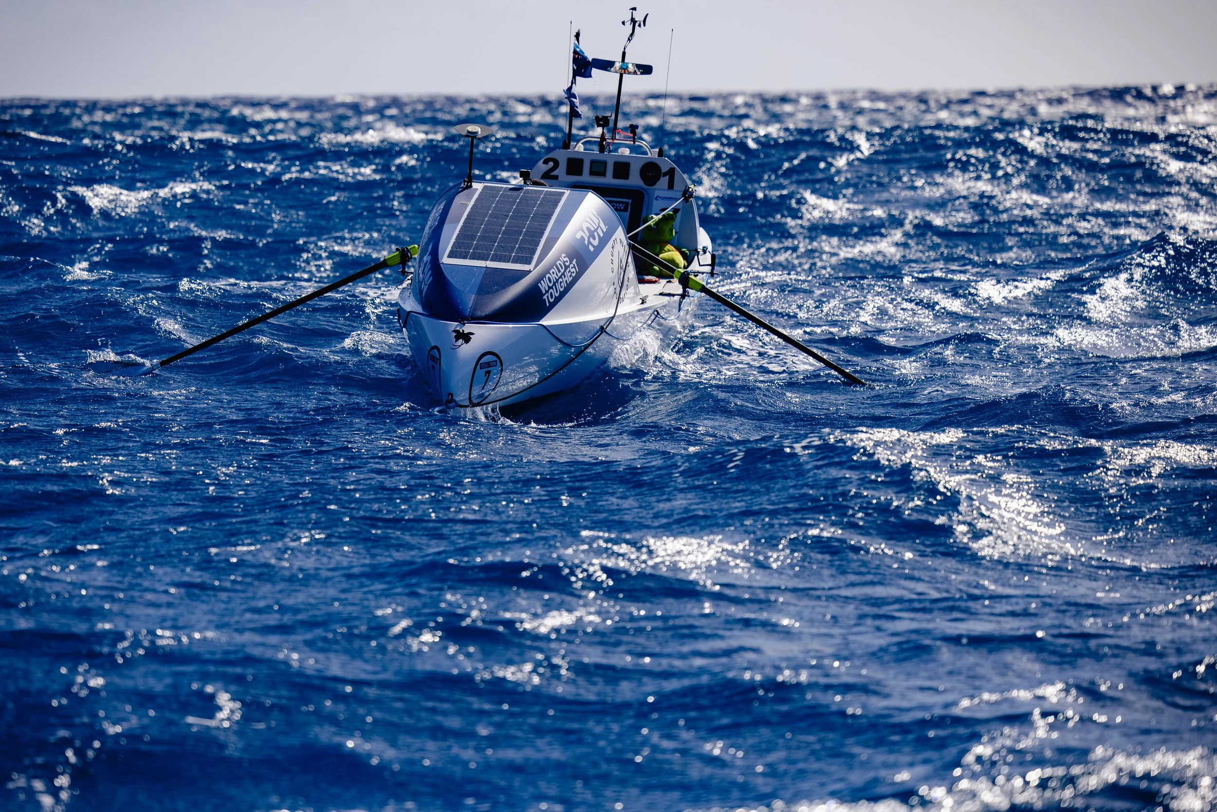 An ocean rowing boat with solar panels on its front is moving through choppy ocean waters. The boat is directed towards the viewer, with the ocean and a cloudy sky in the background.