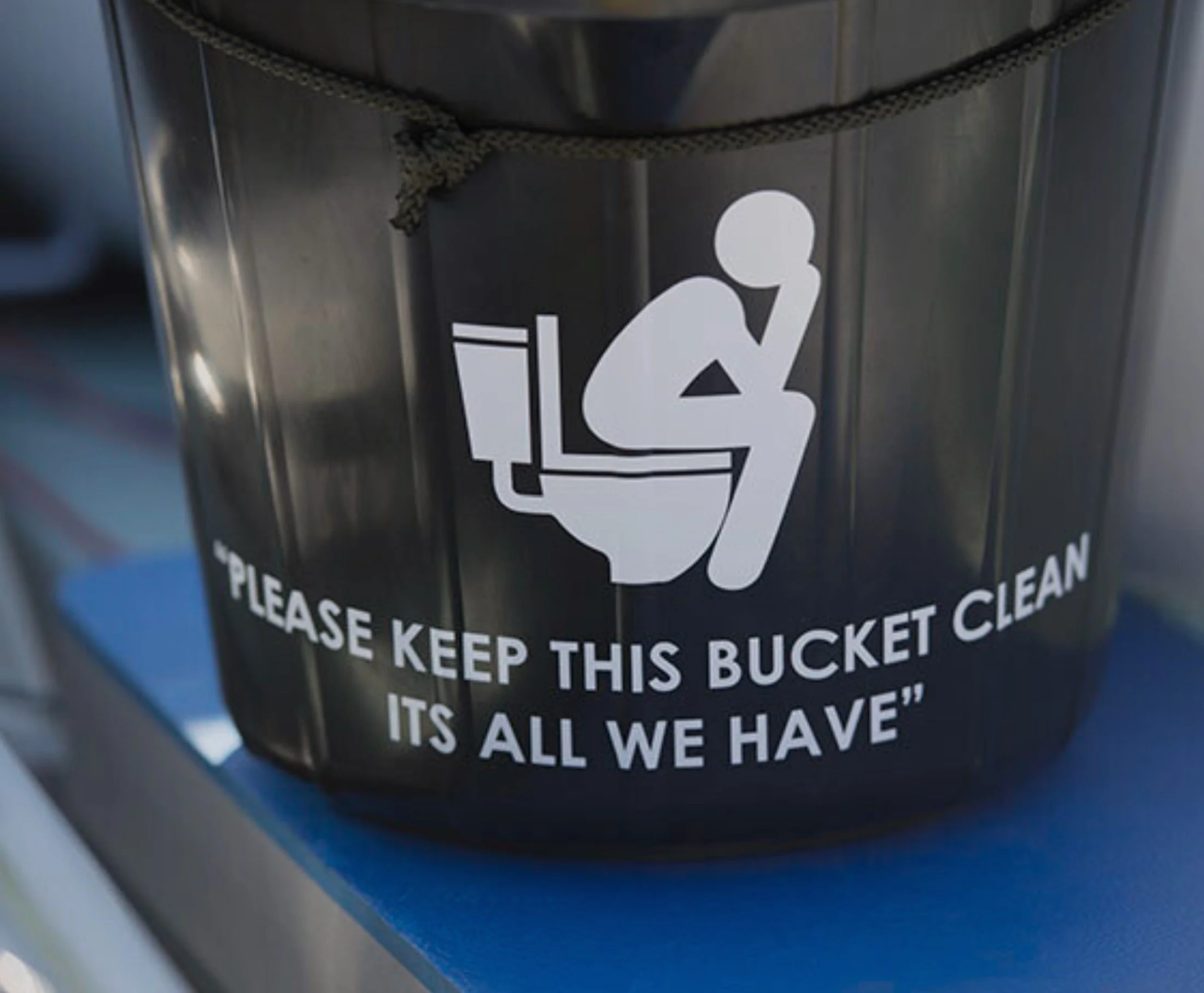 A black bucket with a white graphic of a person sitting on a toilet and reading, along with the text 'Please keep this bucket clean, it's all we have.'