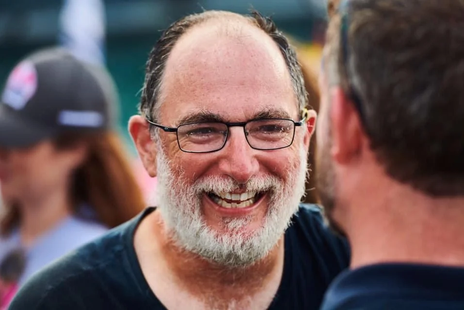 Guy Dresser with a beard and glasses talking to another person outdoors, with a World's Toughest Row crowd around them.