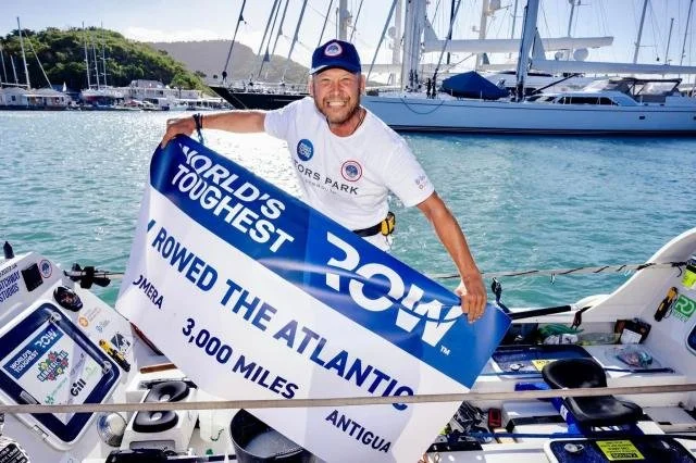 A man smiling on a boat holding a large blue and white banner with the text 'World's Toughest' and 'Rowed the Atlantic 3,000 miles'.