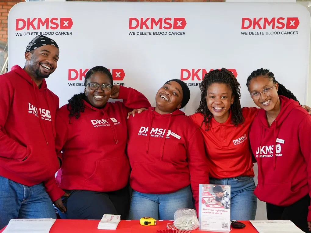 Group of five smiling volunteers at a blood cancer awareness event wearing red hoodies with the DKMS logo, standing in front of a white banner with DKMS branding.