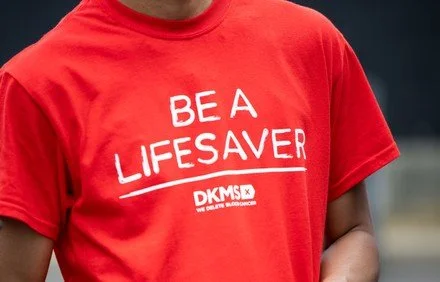 Person wearing a red t-shirt with white text that reads 'BE A LIFESAVER' and a smaller logo 'DKMS' beneath it.