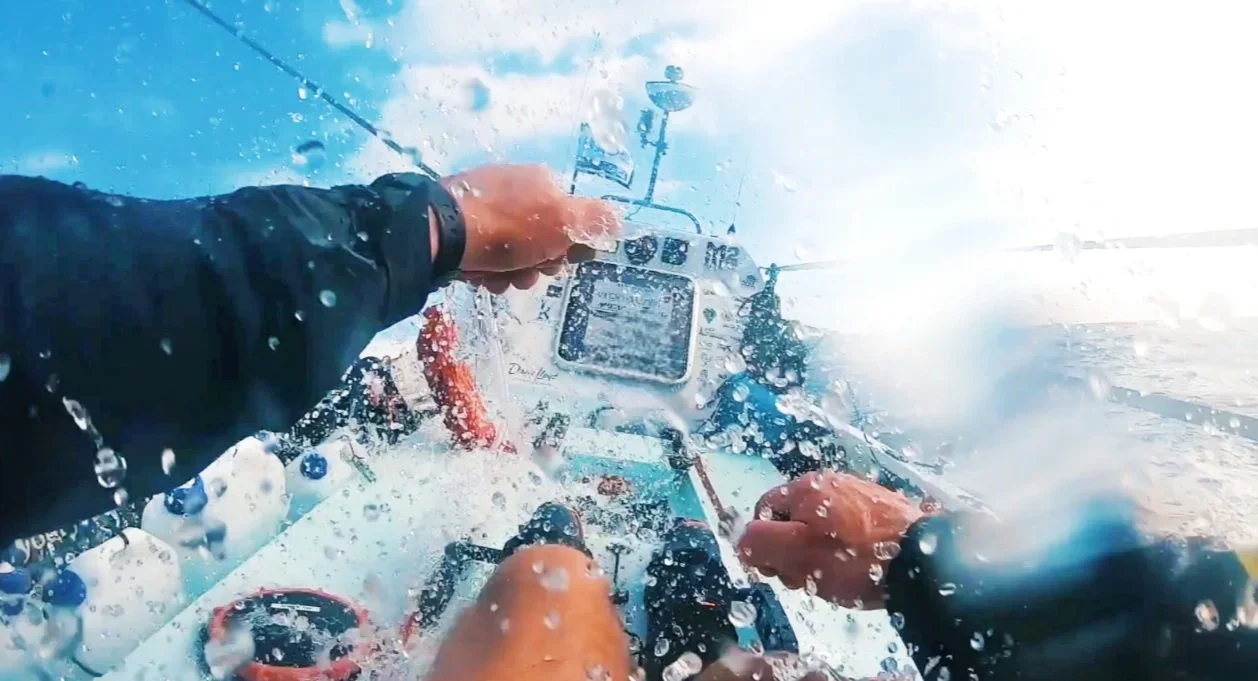 A point of view photo of a man rowing the Atlantic, waves and water are splashing across his arms and legs as he rows in difficult conditions.