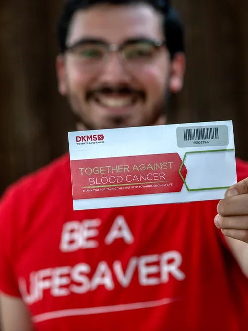 Smiling man wearing glasses holding a card that reads 'Together Against Blood Cancer' and 'Thank you for taking the first step towards saving a life.' He wears a red shirt with partially visible white text.