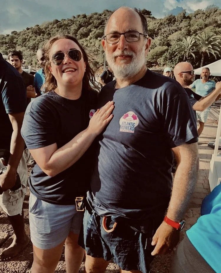 Guy Dresser and his wife, both wearing Atlantic Power shirts, standing outdoors with a group of people and palm trees in the background.