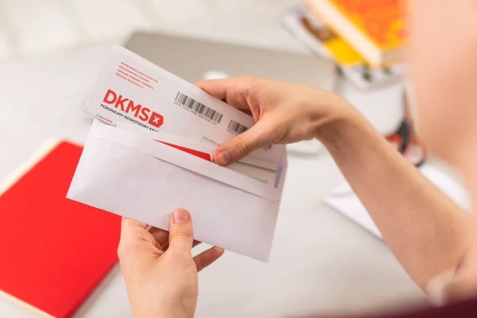 Person holding a white envelope with the DKMS logo, likely containing a health or donation-related document, on a white table with a red notebook and other papers in the background.