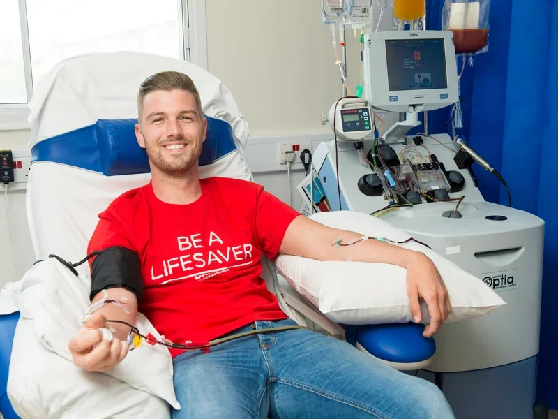 Young man in a hospital bed donating blood, smiling, with medical equipment nearby.