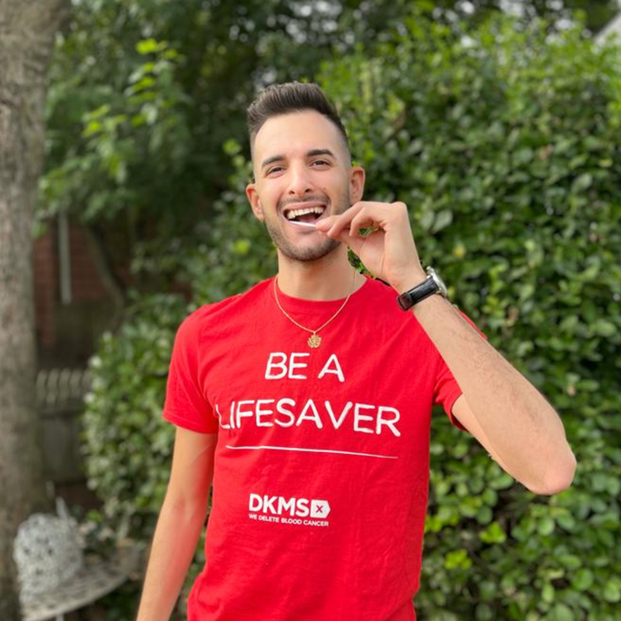 Smiling man wearing a red T-shirt with white text that reads 'Be a Lifesaver' and a blood donation logo, standing outdoors with green bushes in the background.