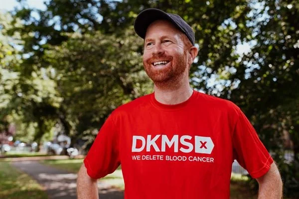 A man with red hair and a beard smiling outdoors, wearing a red t-shirt with white text that says 'DKMS' and 'We delete blood cancer,' and a dark baseball cap.