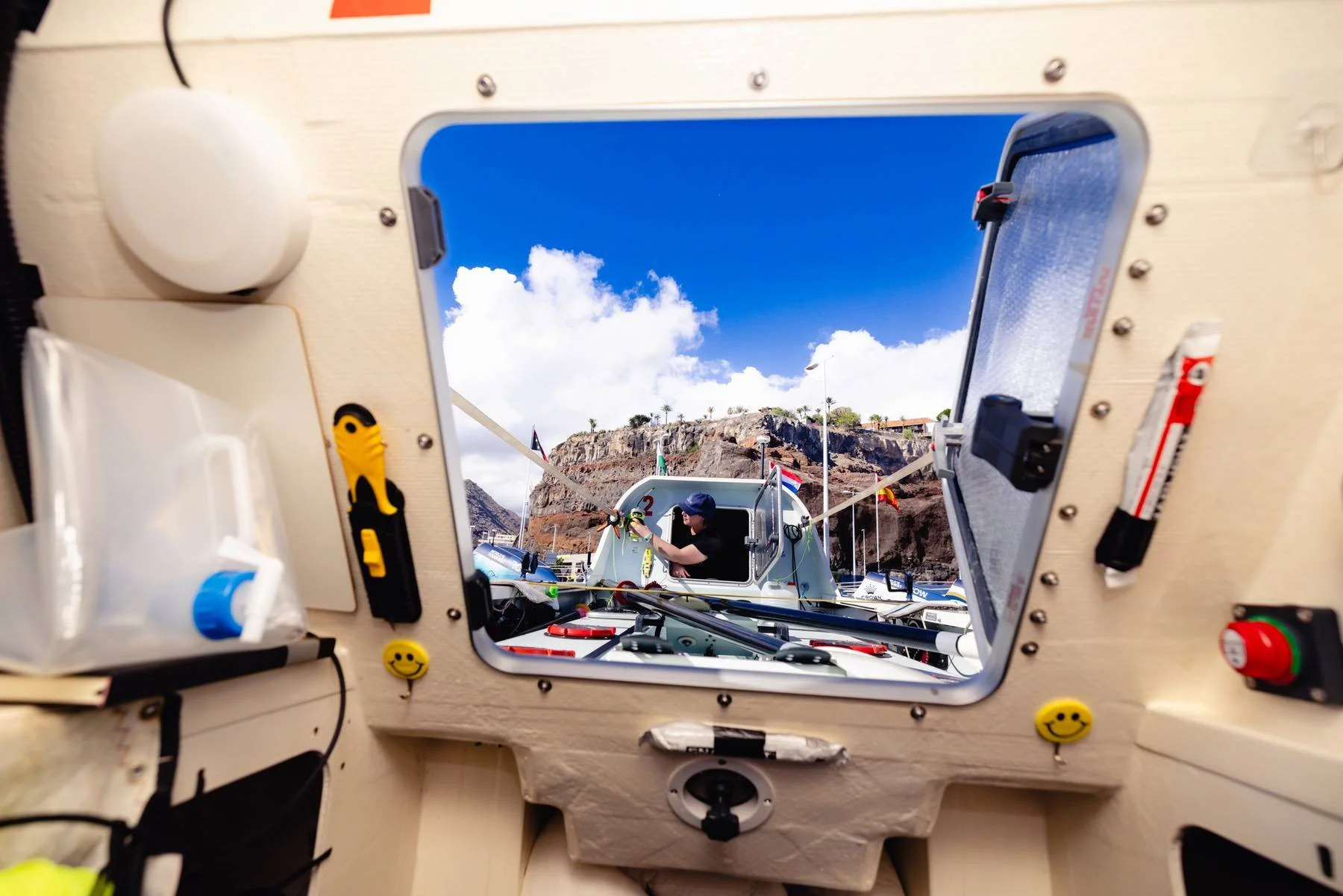 View through an ocean rowing boat hatch showing a person in a small boat on the water with a rocky hill and blue sky in the background.