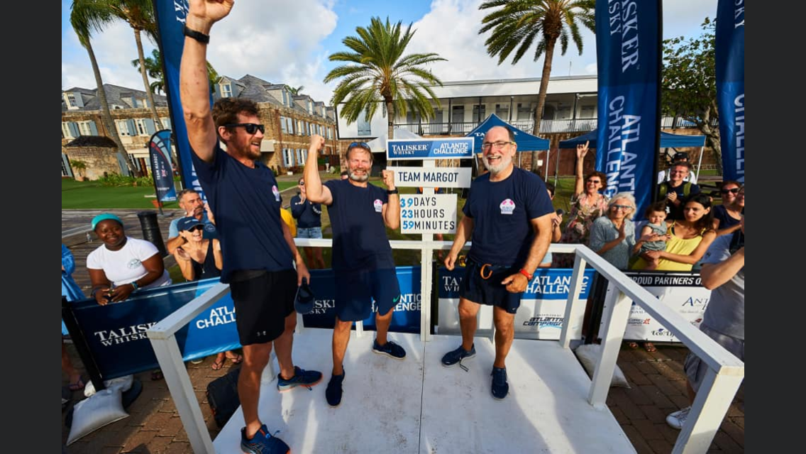 Team Margot standing on a podium celebrating completing the World's Toughest Row, with a crowd of spectators behind them, palm trees, and a blue sky in the background.