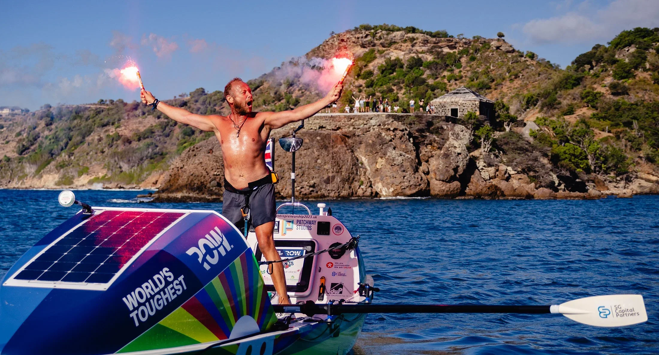 Sam Glover celebrating with flares on a solar-powered ocean rowing boat in the water near a rocky coastline in Antigua.