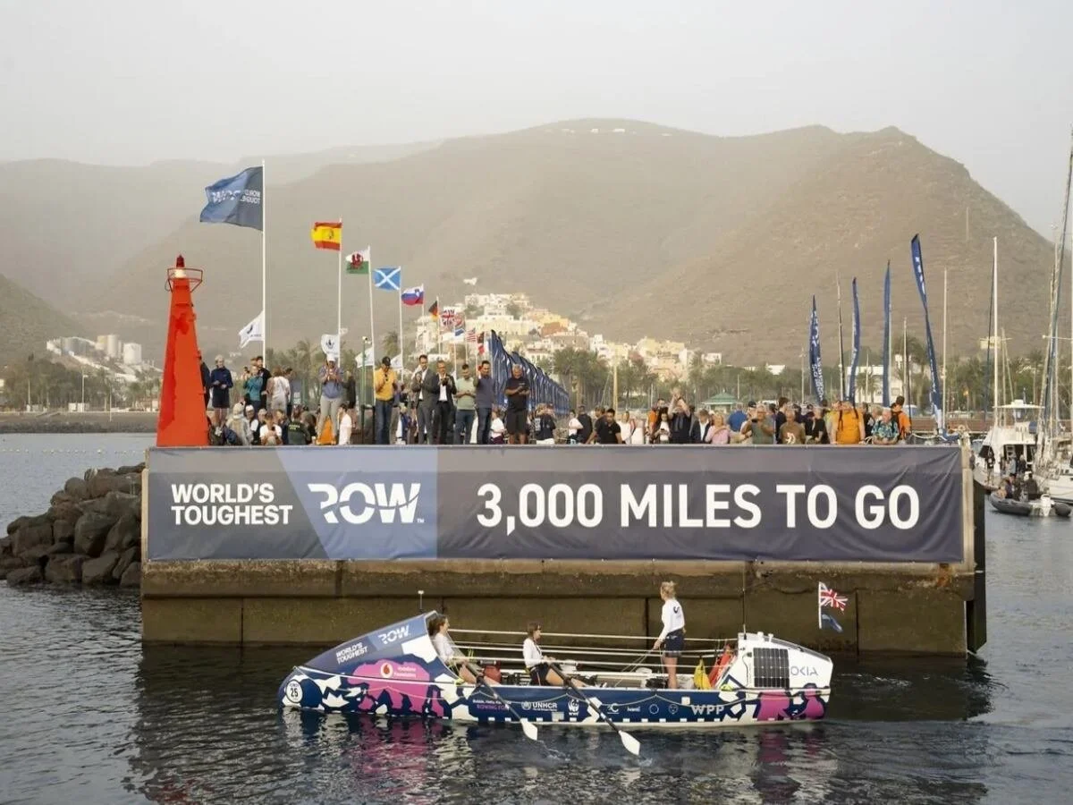 A boat participating in the "WORLD'S TOUGHEST ROW 3,000 MILES TO GO". People are gathered on a pier with flags from various countries and on a boat, and a scenic hillside is visible in the background.