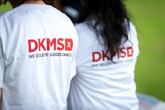 Two people wearing white T-shirts with red and black text promoting blood donation for DKMS, a blood cancer charity, with a green background.
