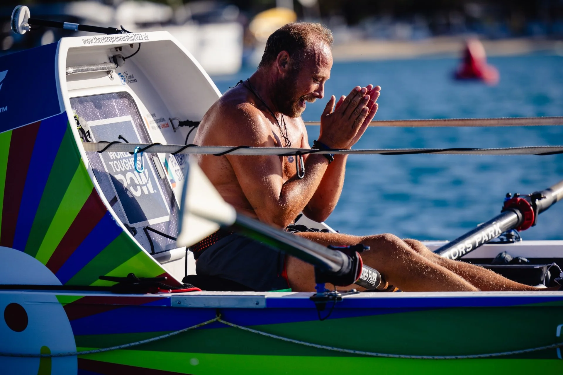 Sam Glover sitting in a boat with his hands pressed together in a prayer-like gesture, smiling, after completing the World's Toughest Row