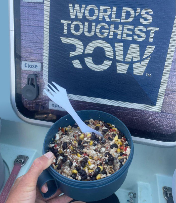 A bowl of food, held by a person on an ocean rowing boat with a sign reading 'World's Toughest Row' in the background.