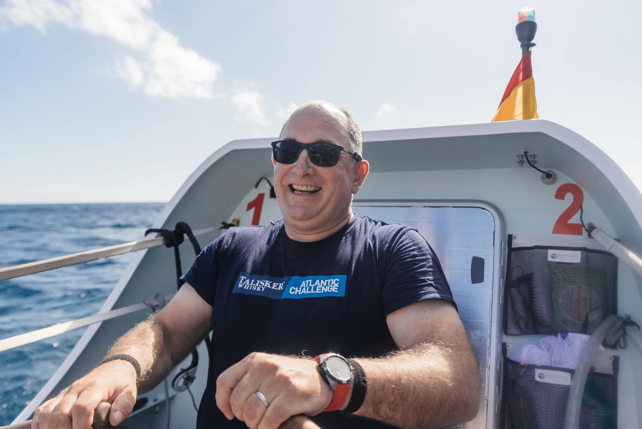 Guy Dresser smiling and wearing sunglasses rowing the Atlantic in Team Margot, wearing a black T-shirt with the words "Talisker Whisky Atlantic Challenge" on it.
