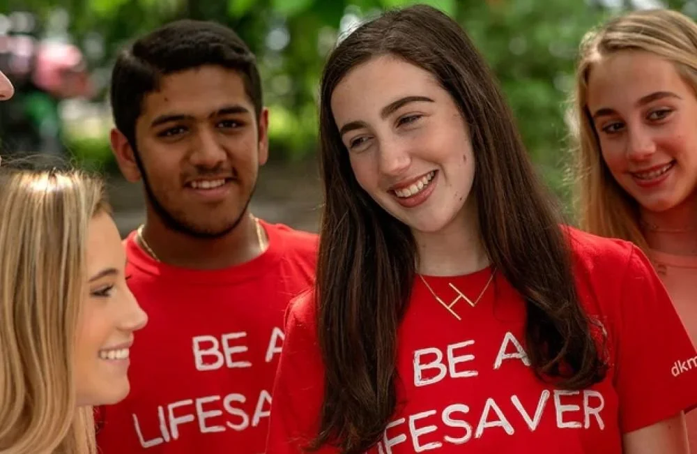 Group of smiling young people outdoors, wearing red shirts that say 'Be a Lifesaver'.