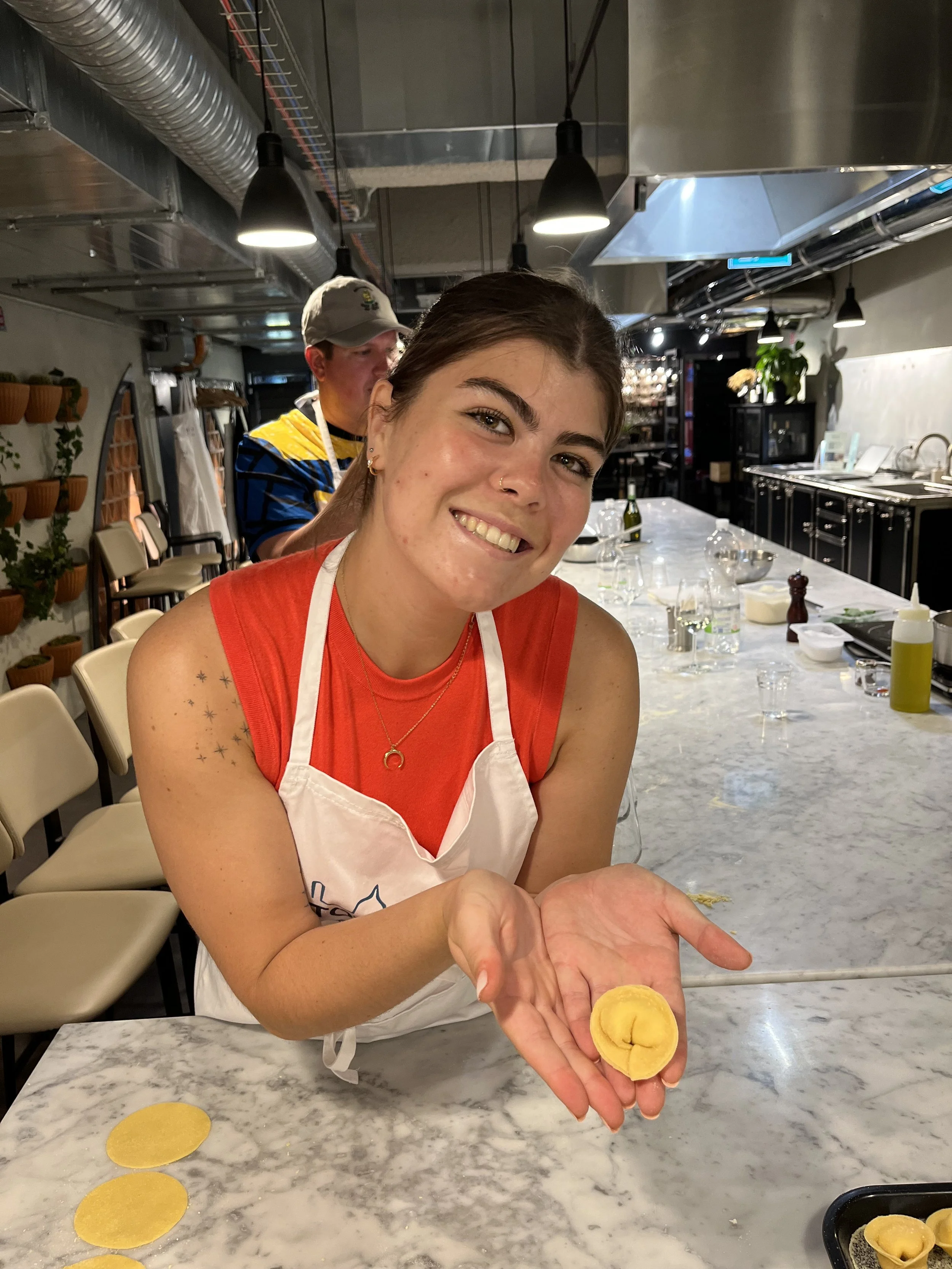 A young woman smiling and showing a yellow dumpling she made, in a kitchen or bakery setting with a marble counter, cooking utensils, and a man in the background.