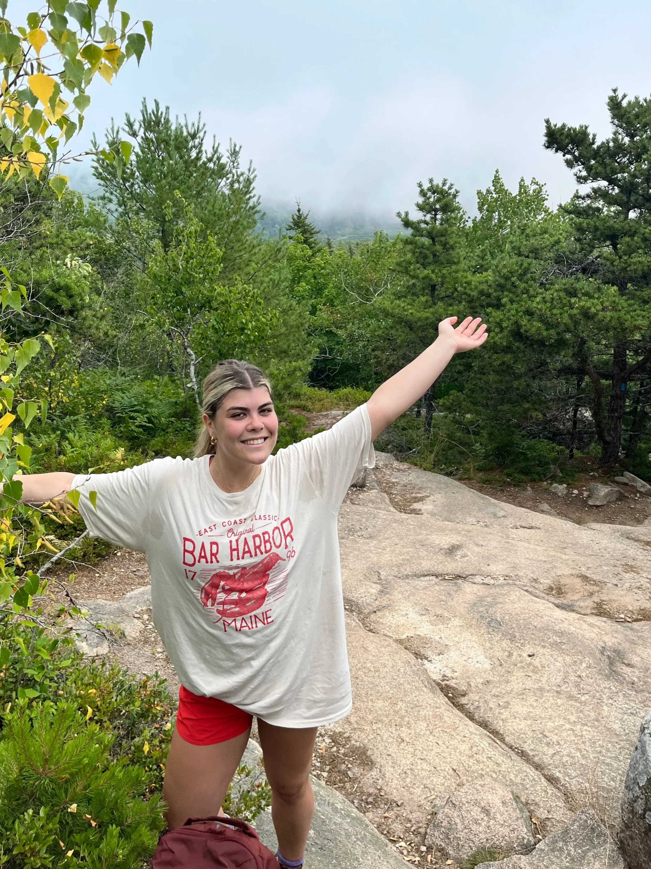 A young woman smiling with arms outstretched standing on a rocky trail in a lush green forest with foggy mountains in the background.