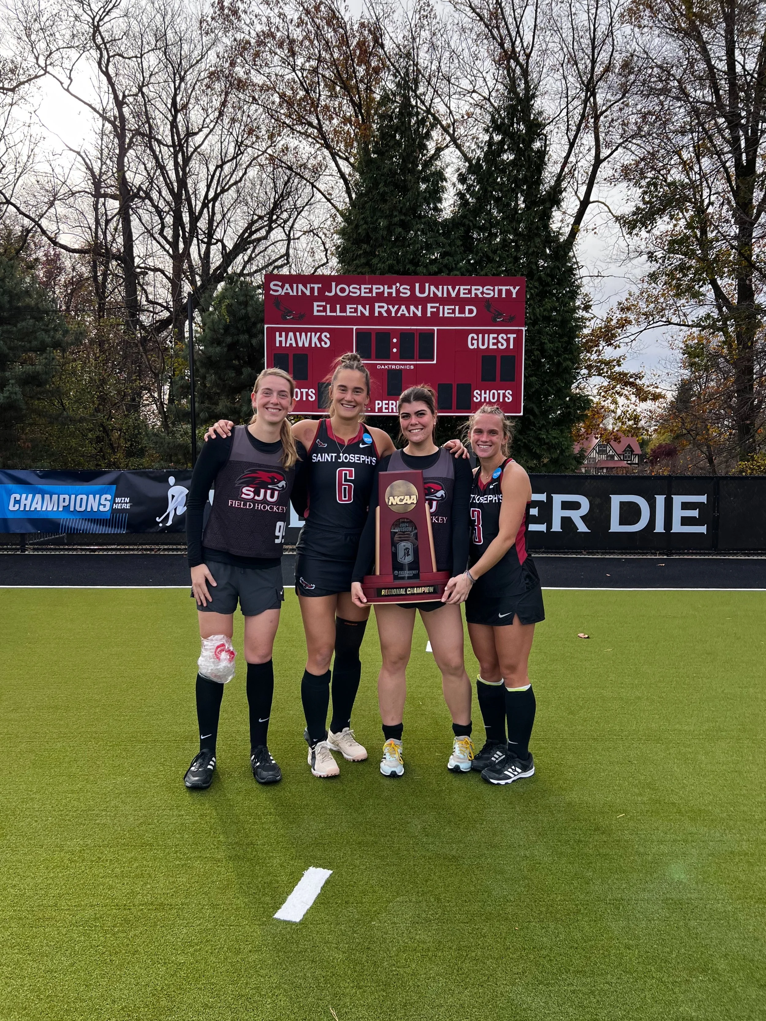 Four female college field hockey players in black and red uniforms celebrating with a regional championship trophy on a grass field, with a scoreboard and trees in the background.