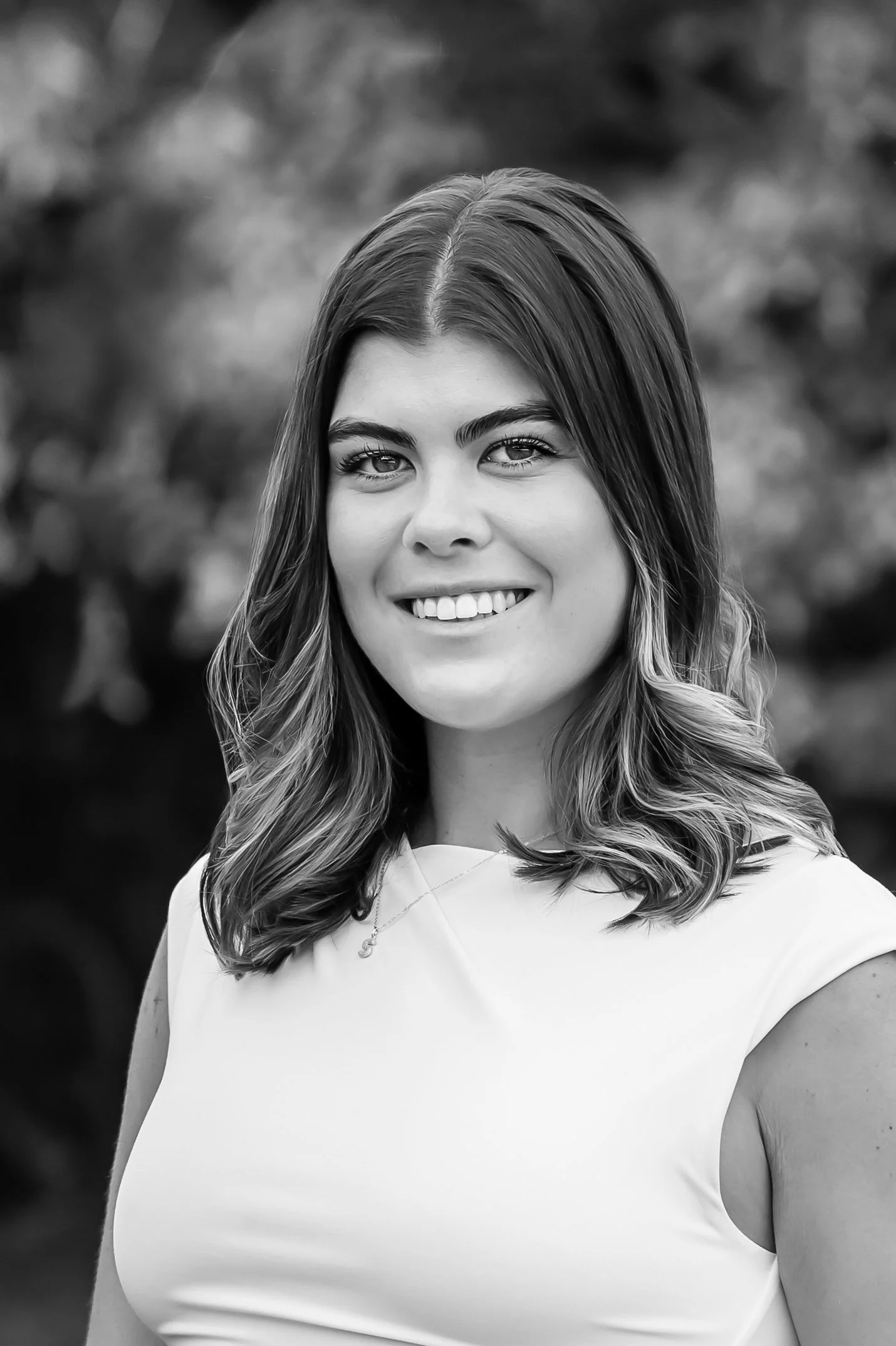 A black and white portrait of a young woman with shoulder-length wavy hair, smiling, wearing a sleeveless top and a necklace, with a blurred outdoor background.