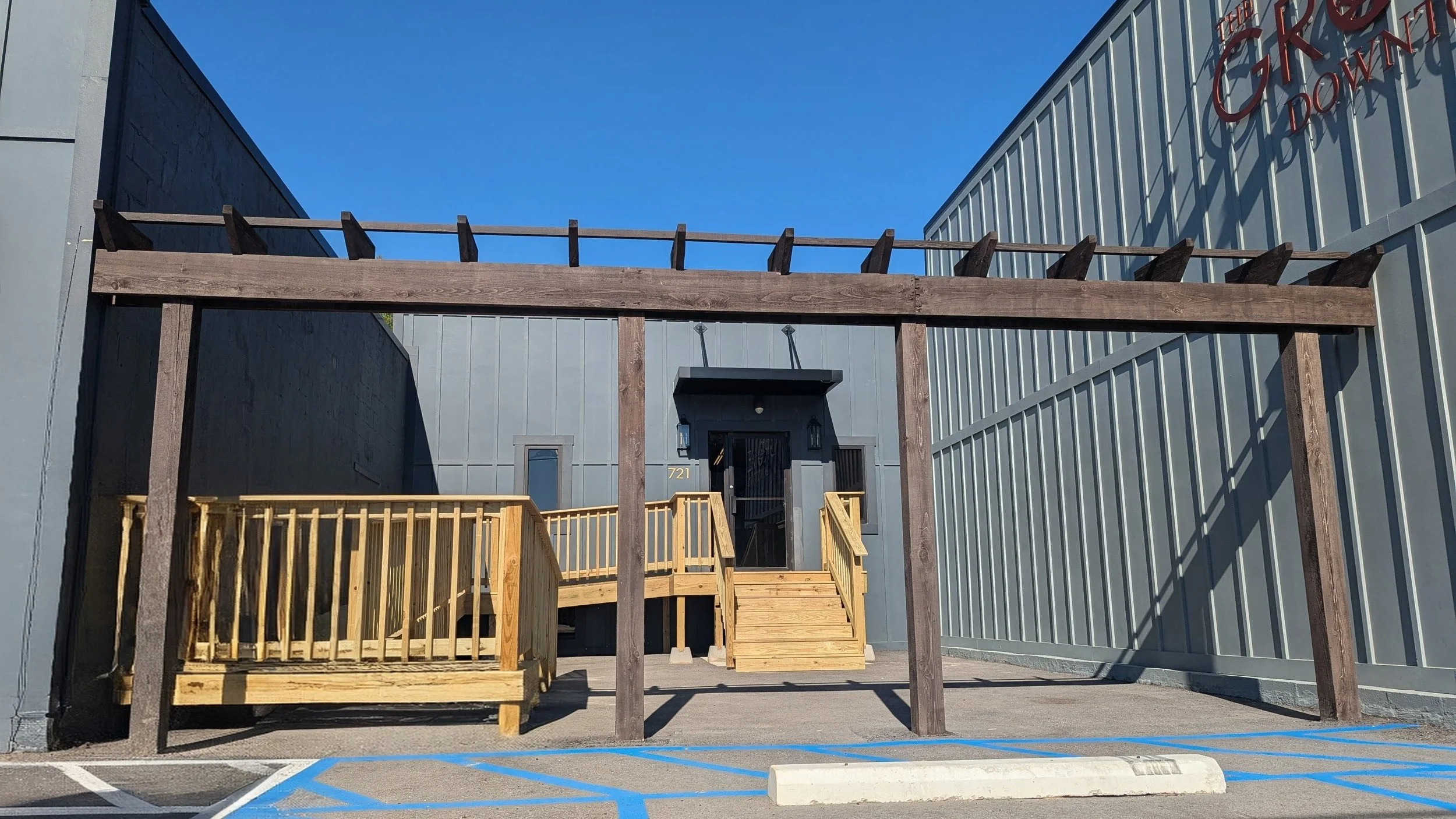 A no parking zone marked with blue lines in front of a building with a wooden frame that looks like it might support an overhead element. The building has a stairway leading to a black door with the number 721, and the background features a gray industrial exterior with red GP logo and part of a gray metal fence on the right. The sky is clear and blue.