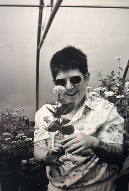 A person wearing sunglasses and a patterned shirt smiling while holding a flower inside a greenhouse or plant nursery.