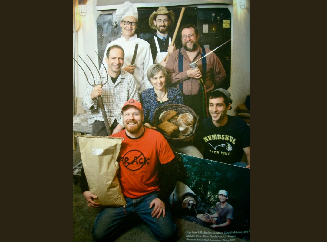 Group of seven people posing together holding farming tools and bread, around a table with a basket of bread, in a cozy indoor setting.