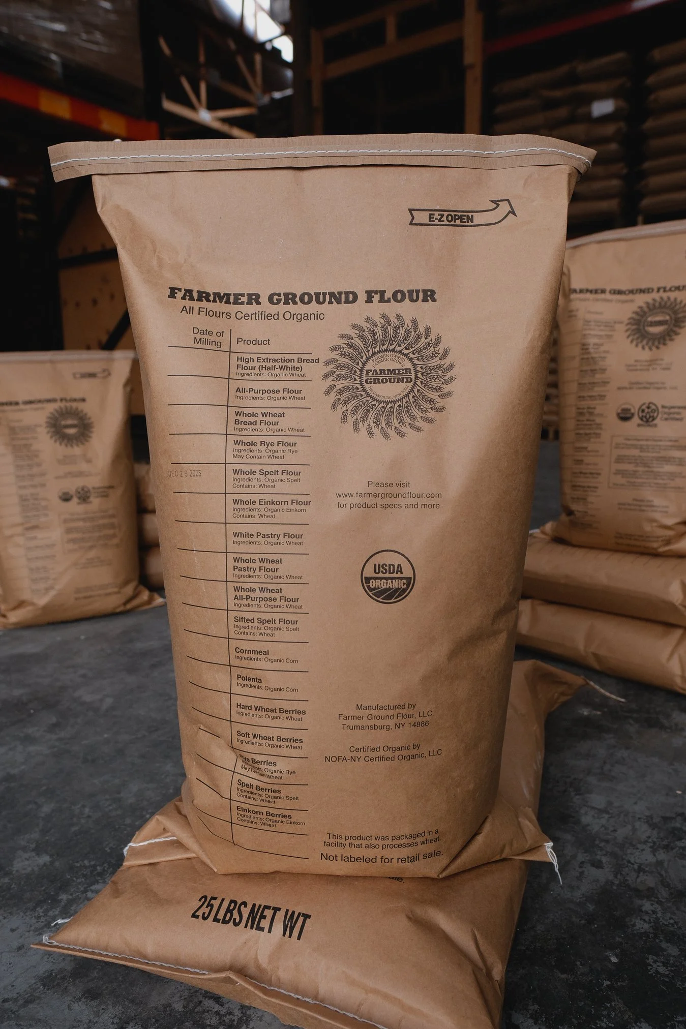 Brown paper bag of Farmer Ground Flour, 25 pounds, with product list and USDA Organic label, placed on a concrete floor with other similar bags in the background.