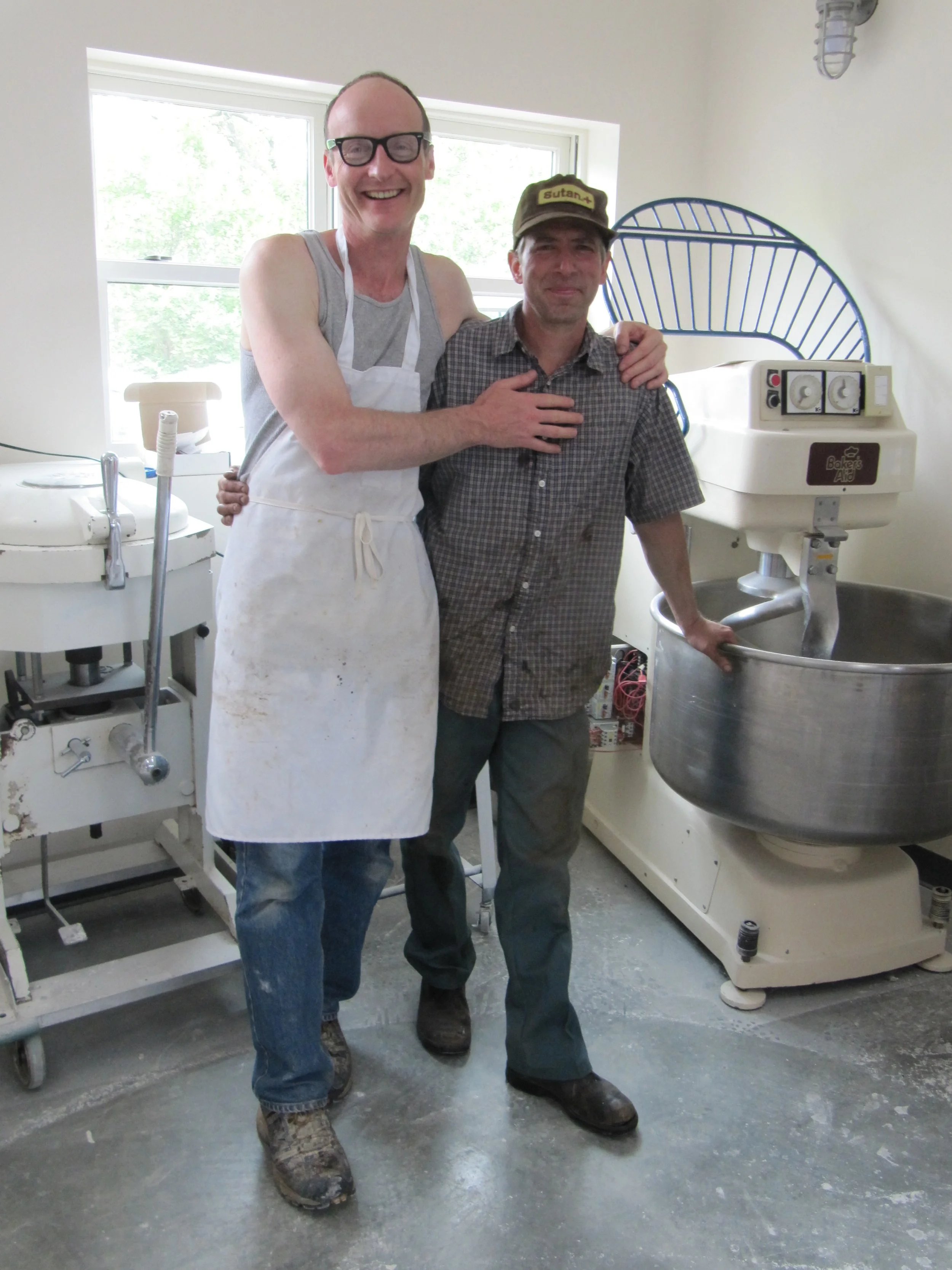 Two men standing together in a bakery, smiling. One man is wearing an apron and glasses, the other is wearing a cap and checkered shirt. Behind them are bakery equipment, including a large stand mixer and another machine, with a bright window in the background.