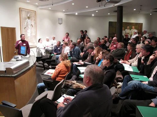 A man is speaking to an audience in a conference room with a mixture of adults seated and listening attentively, some taking notes. The room has artwork on the walls and a few people standing at the back.