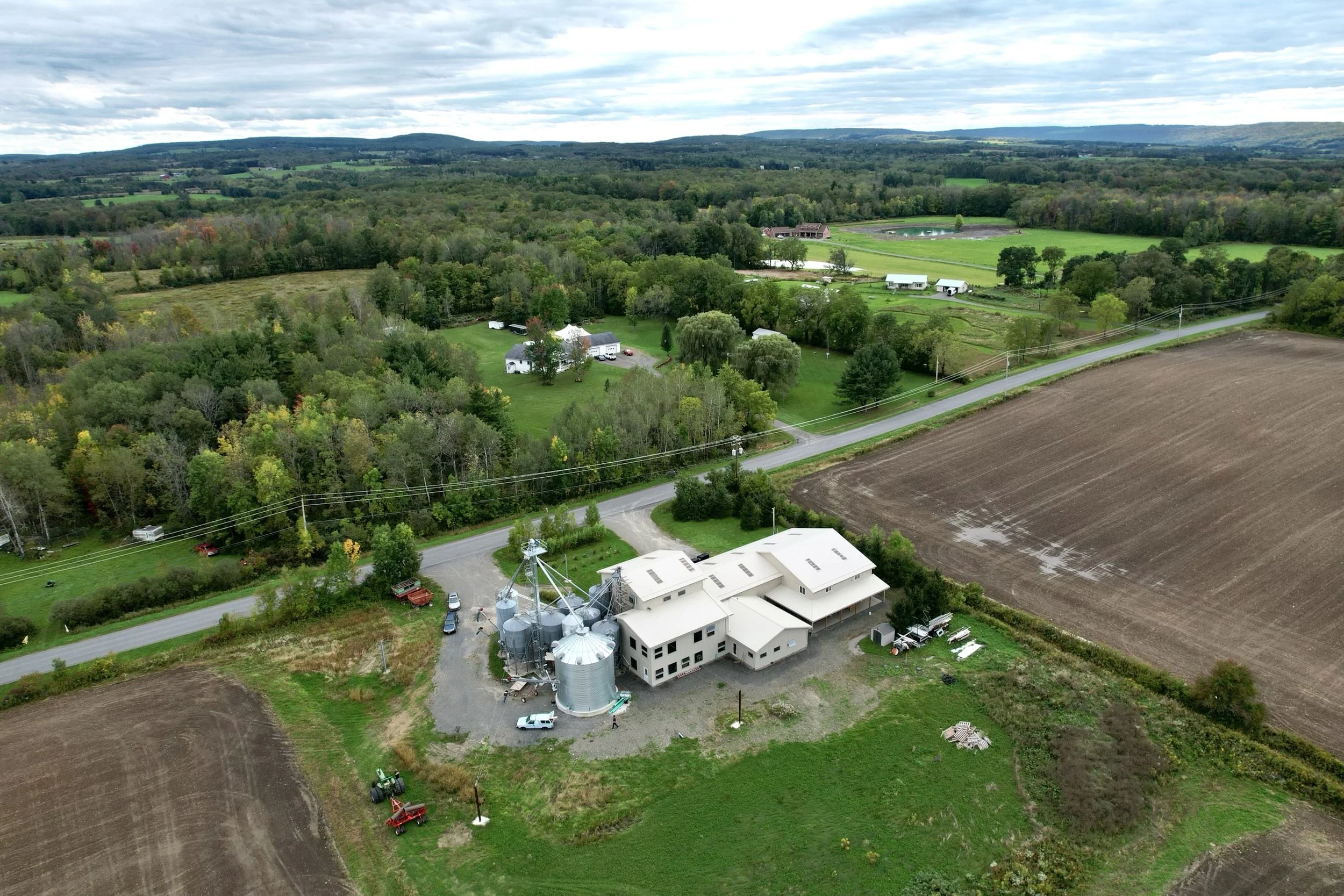 Aerial view of the Farmer Ground Flour Mill location, silos, and green fields, surrounded by scattered trees, roads, and neighboring farmhouses in a rural landscape.