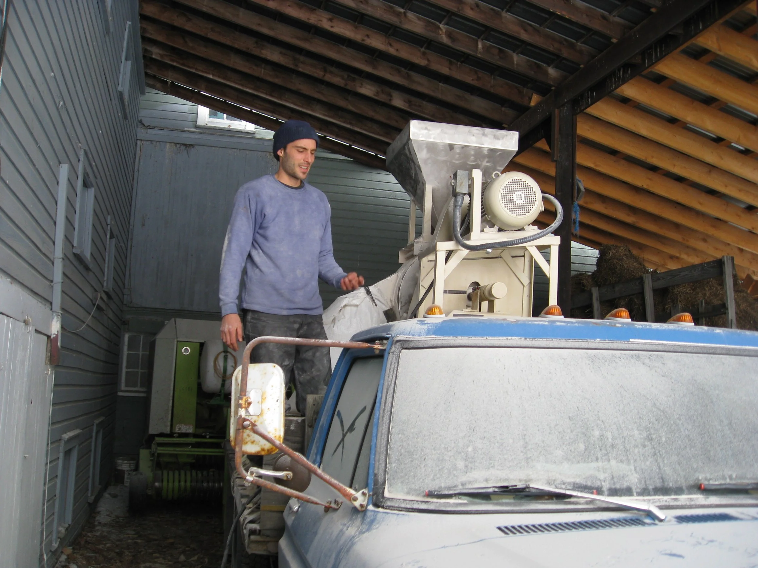 A man standing on the back of a truck with stone mill mounted in the bed of the truck, wearing a beanie and a long sleeve shirt, inside a barn or garage with a wooden ceiling.