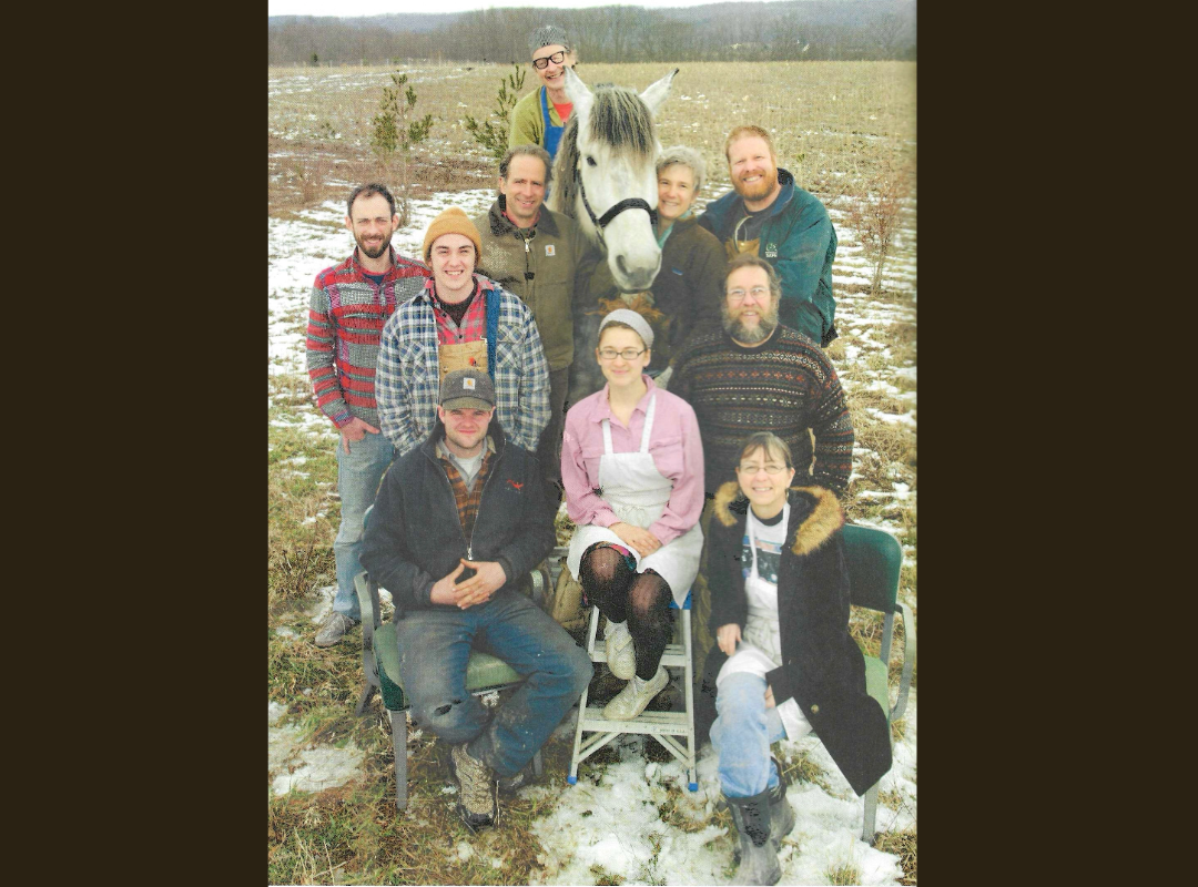 A group of nine people and a white horse in a snowy field, with some small trees and a cloudy sky in the background.