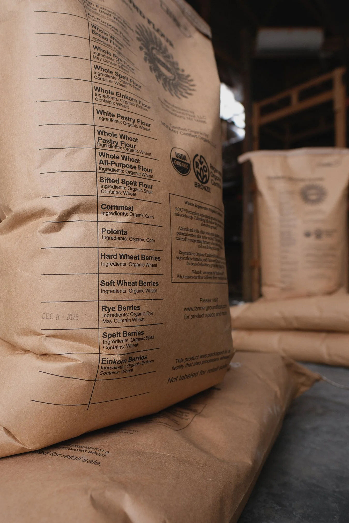 Close-up of a brown paper bag labeled with various types of organic flour and berries, including whole wheat, spelt, einkorn, and rye, with certifications like USDA Organic and Non-GMO Project Verified.