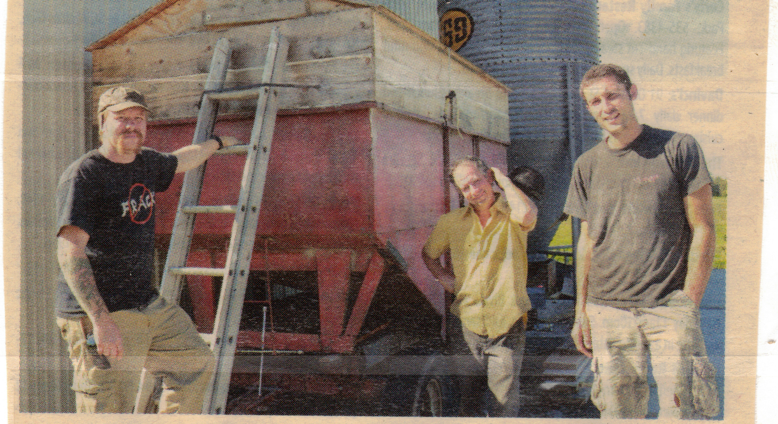 Greg Russo, Thor Oechsner, and Neal Johnston, standing outdoors near a red wooden structure and a metal silo; one man is on a ladder, the other two are standing on the ground, smiling at the camera.