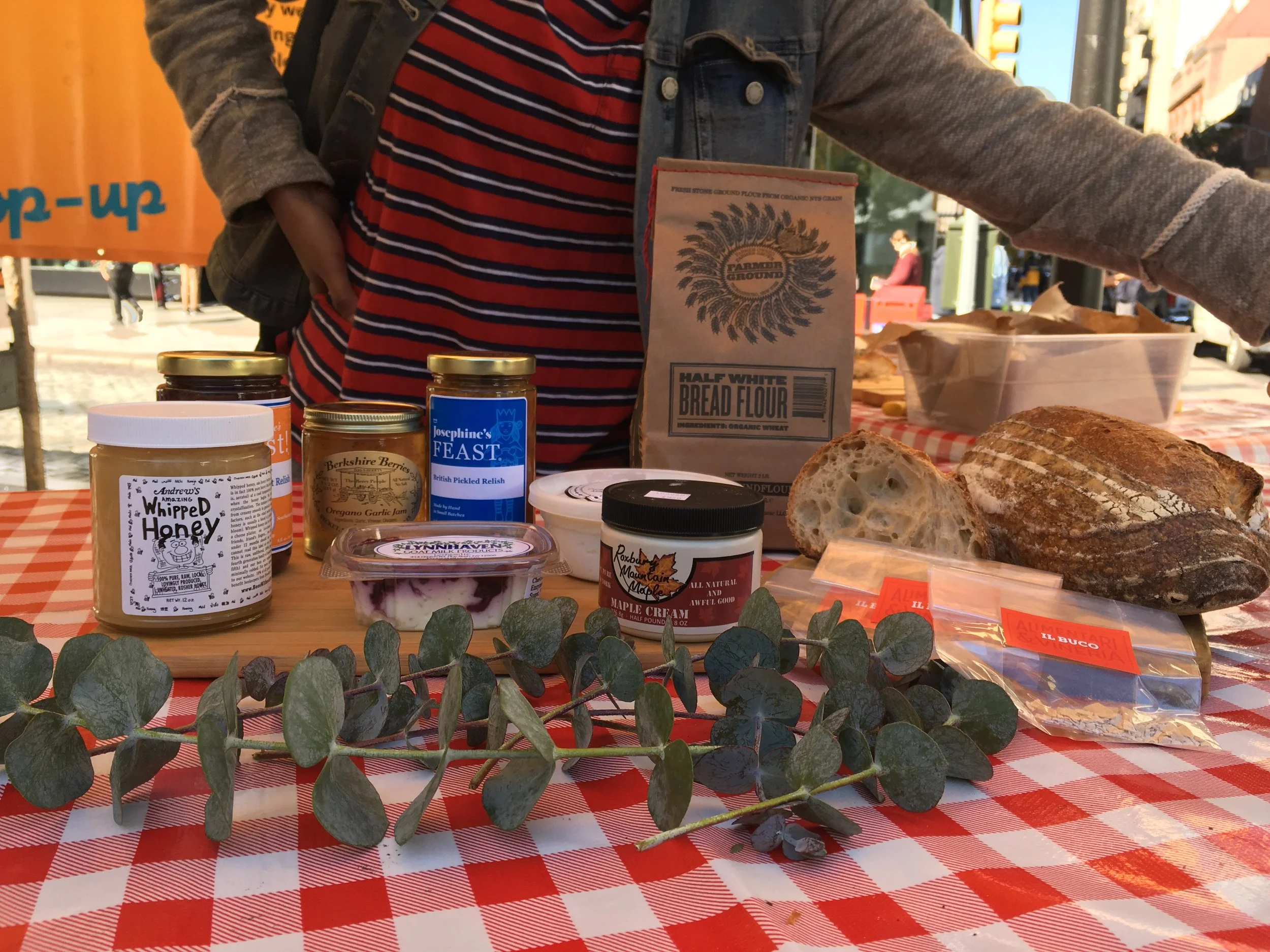 Assorted baked goods and food products on a table with a red-and-white checkered tablecloth, including a loaf of bread, a jar of whipped honey, jars of jam or relish, a container of yogurt or dessert, and a bag of bread flour.
