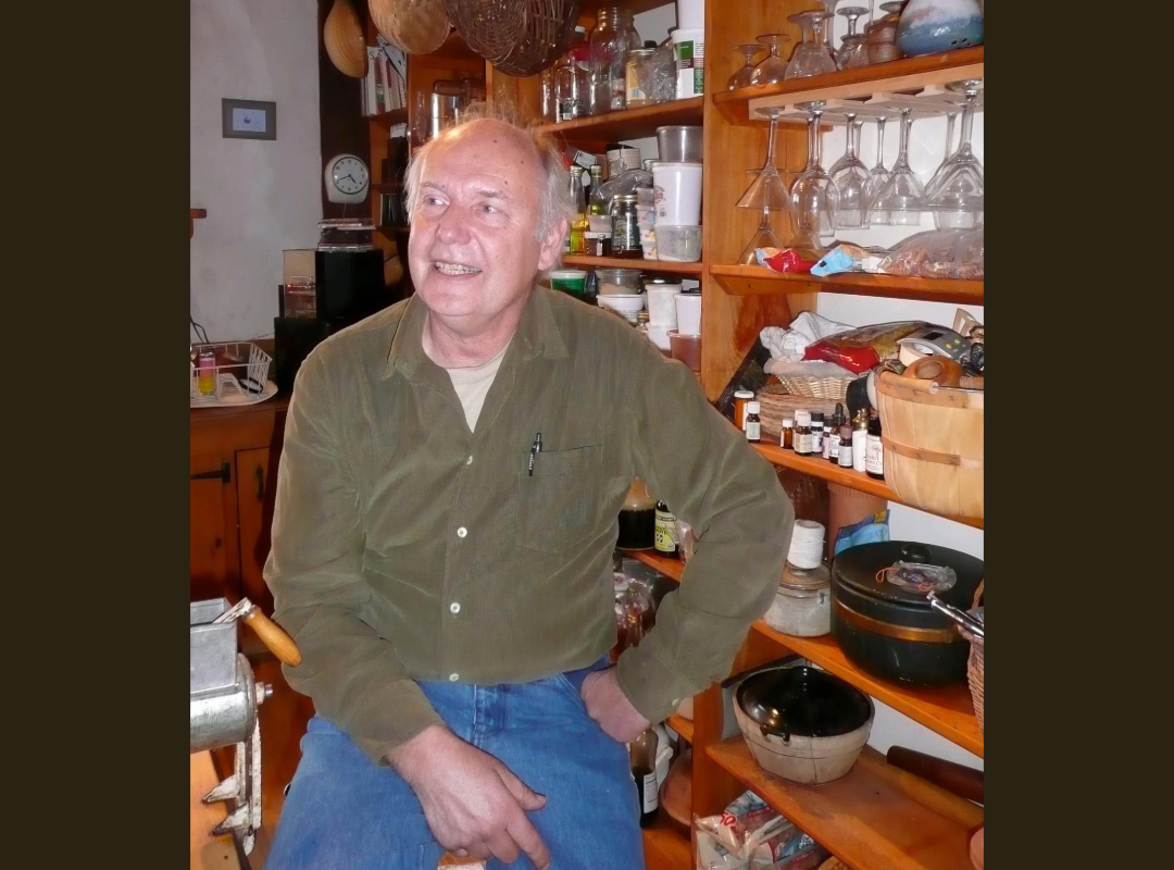 A man with gray hair smiling and sitting in a kitchen surrounded by shelves filled with glasses, bottles, jars, and kitchen items.