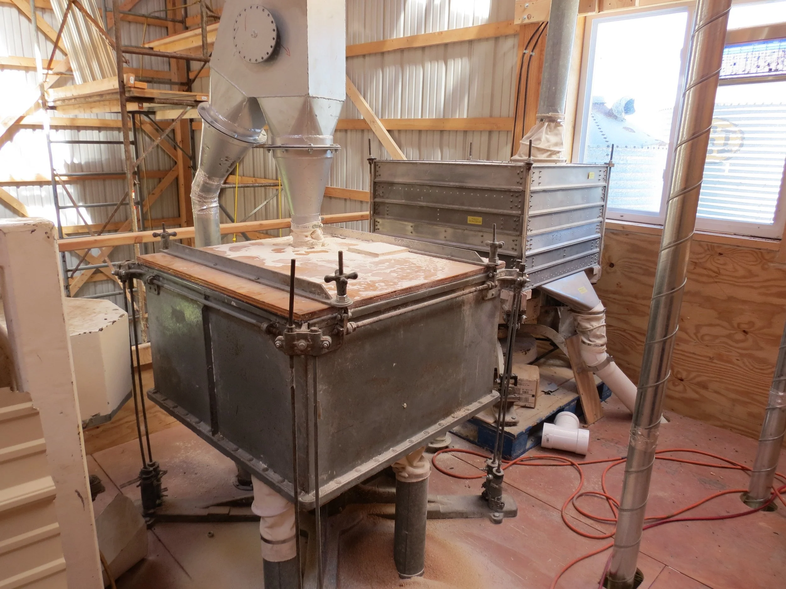 Industrial flour sifter in a sunny room with lots of pipes around it and running to it.