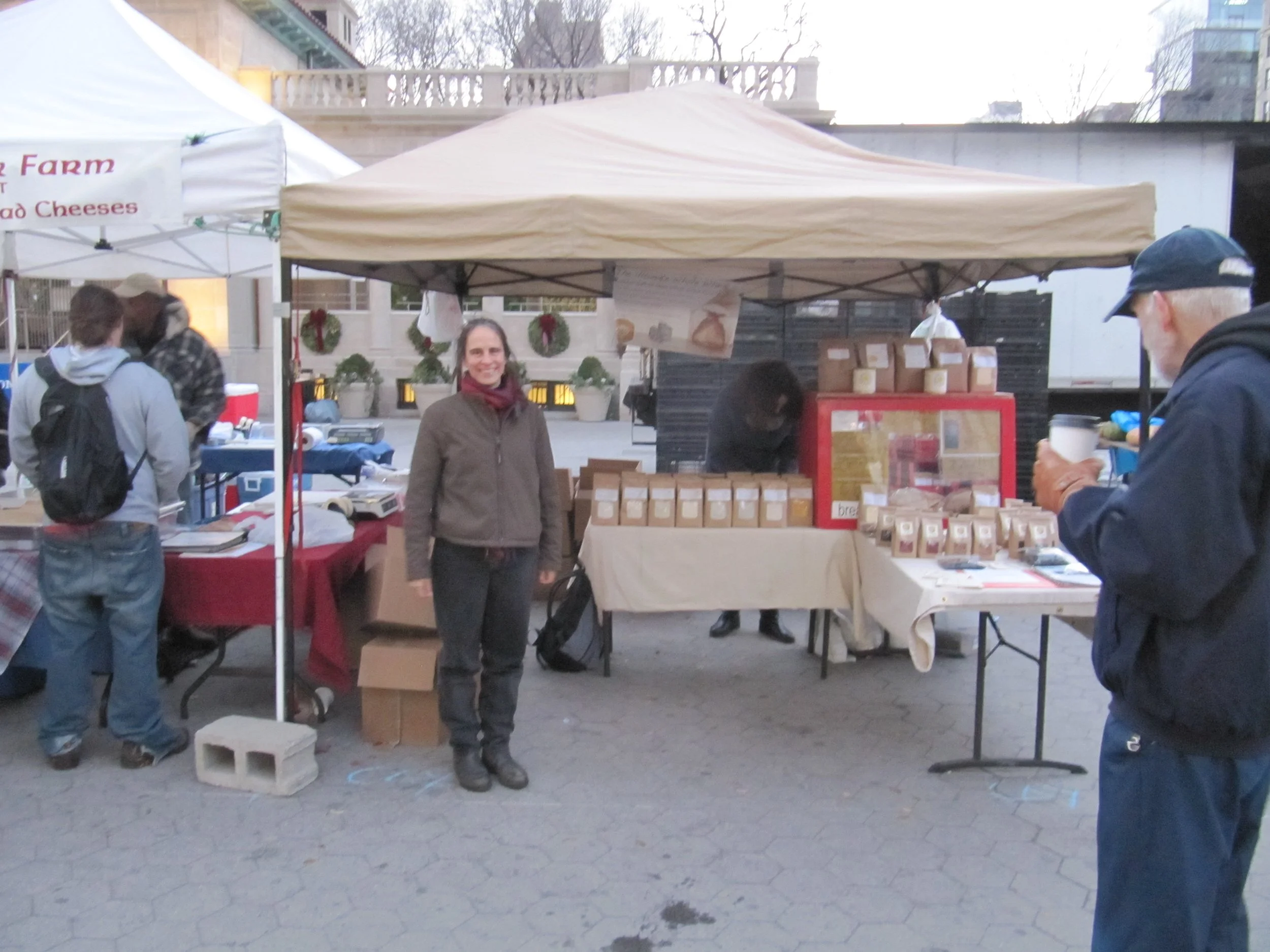 A woman standing in front of a market stall with jars displayed on tables. Other people are shopping and browsing nearby. The scene takes place outdoors on a city street.