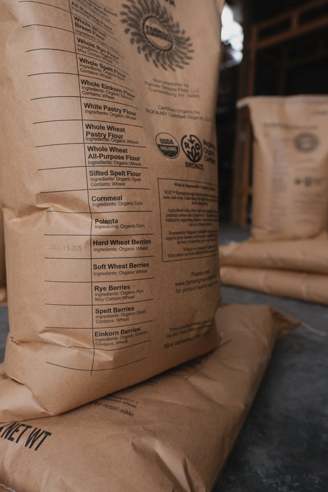 Close-up of a brown paper bag of organic flour containing various types including whole wheat, spelt, einkorn, and rye, with other grain products, with a wooden crate in the background.