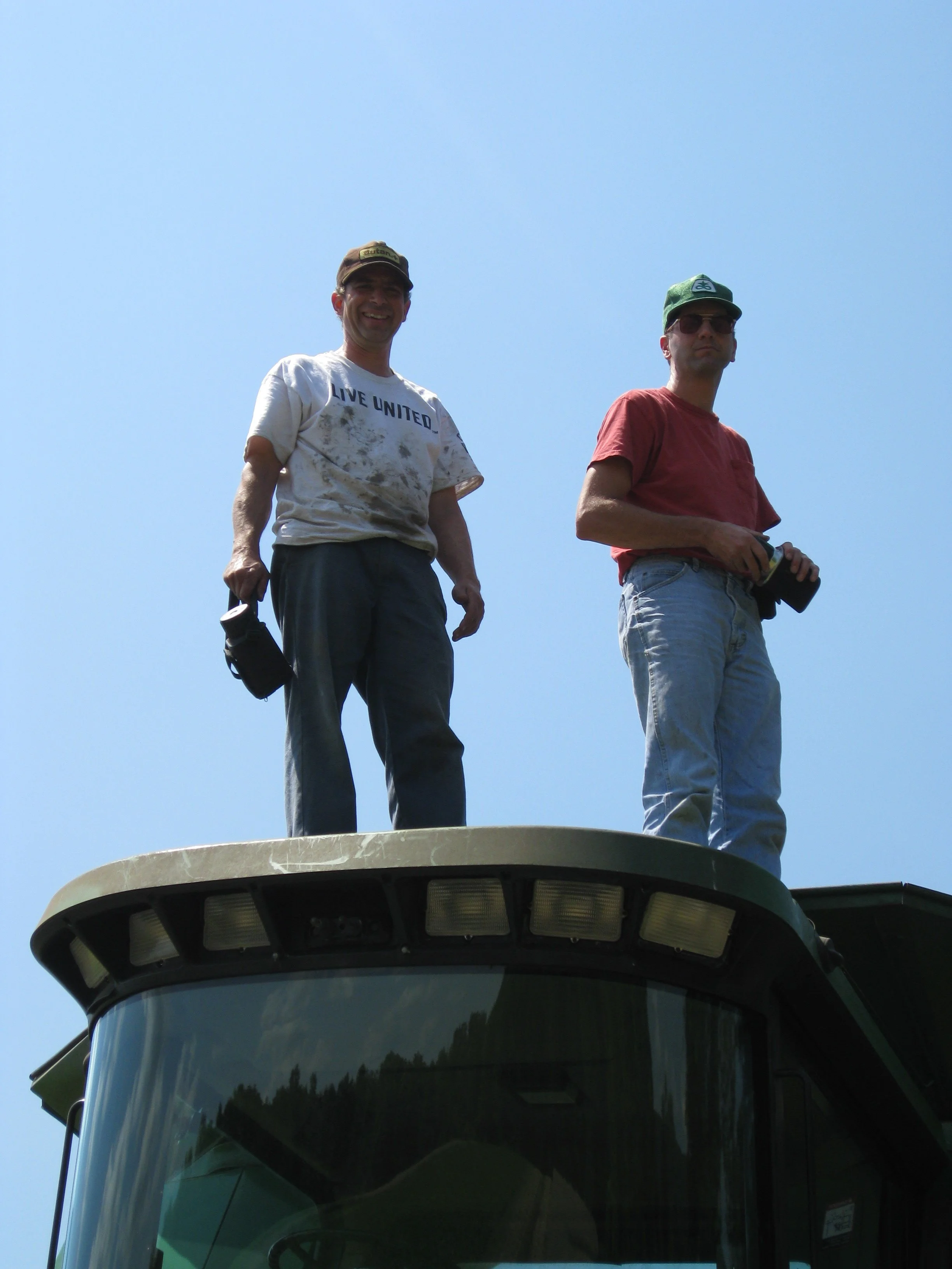 Two men standing on top of a grain storage bin, against a clear blue sky.