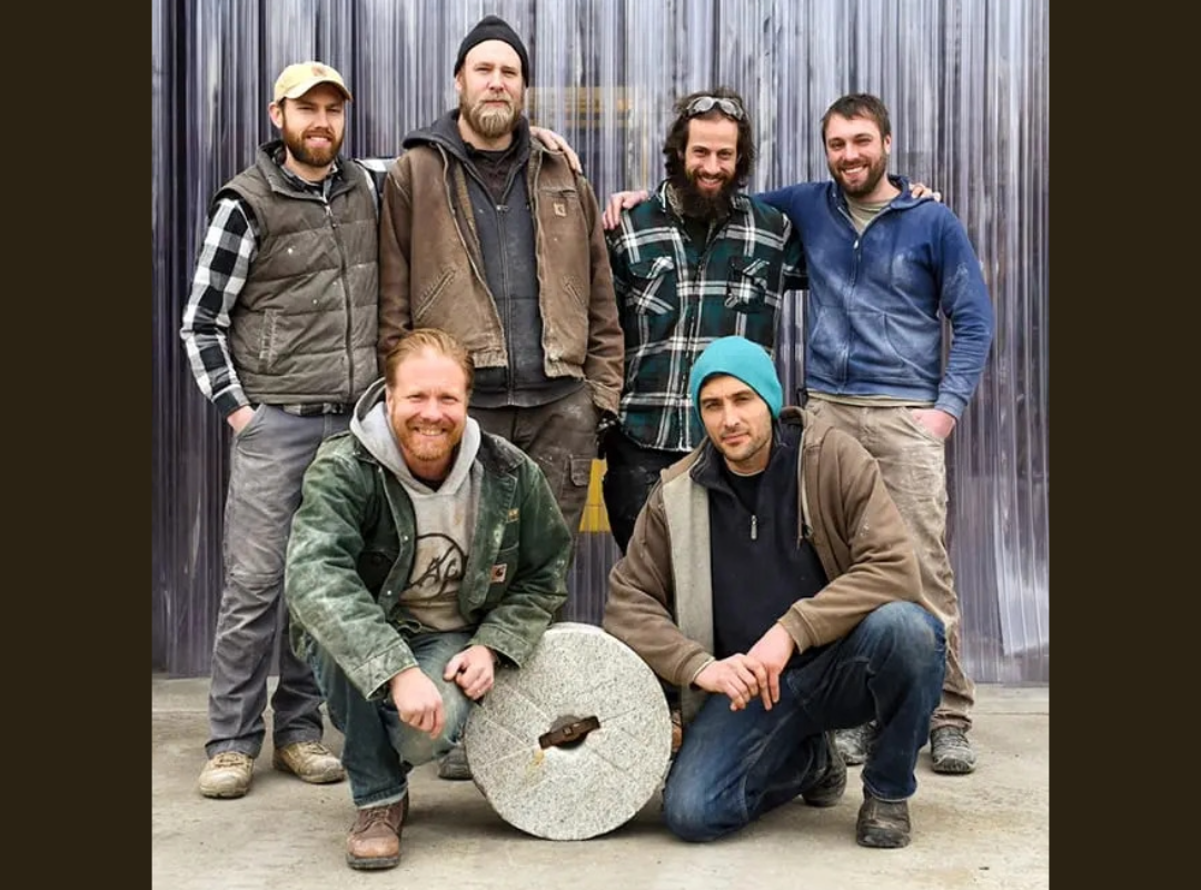 Six men posing outdoors in front of metallic background, two are kneeling, the others standing, some with arms around each other, casual clothing, some wearing jackets and beanies, one with an old millstone staged in front of them on the ground.