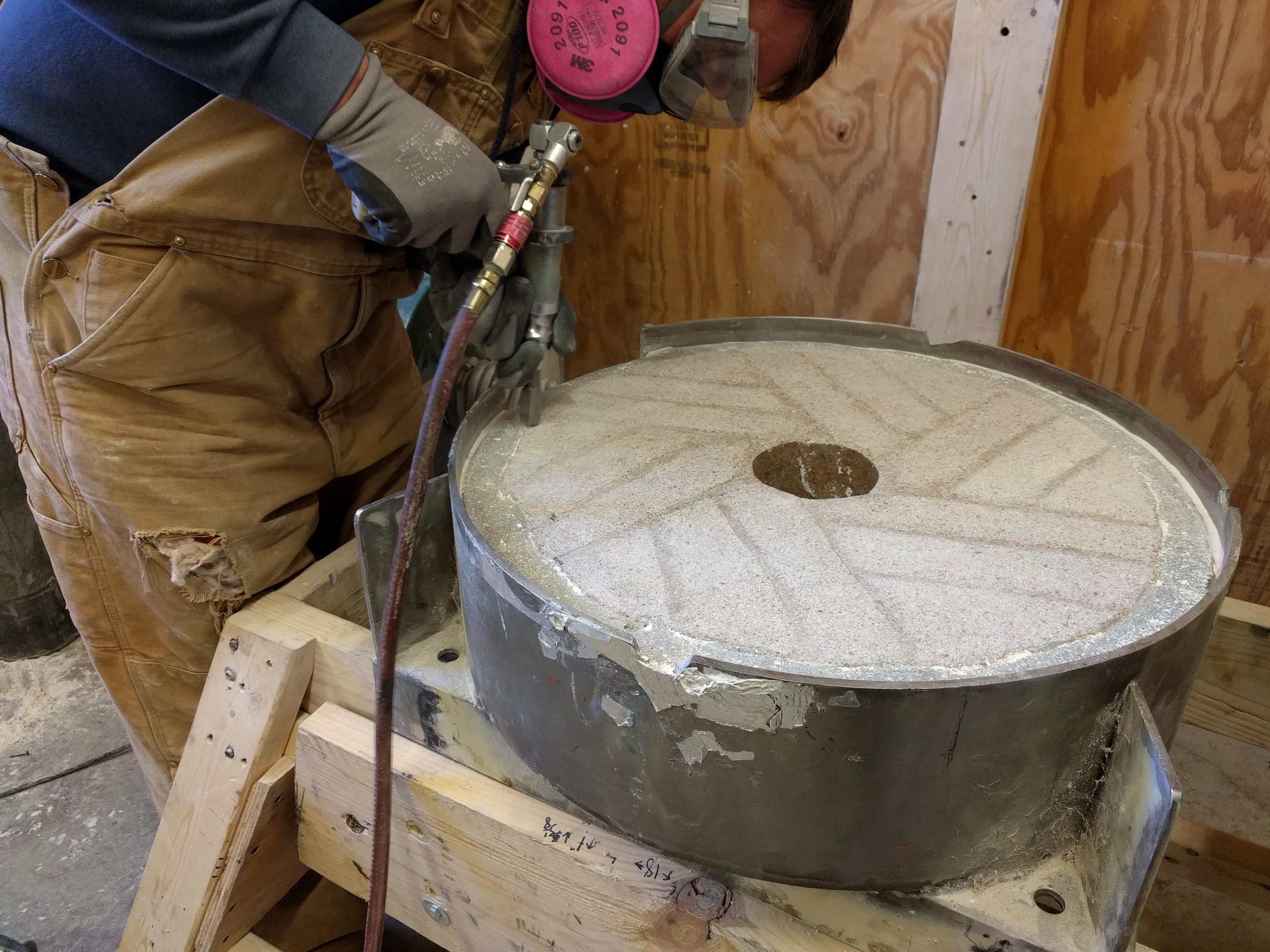 Person using a pneumatic chisel to sharpen a millstone that is laying on some robust saw horses or a custom bench.