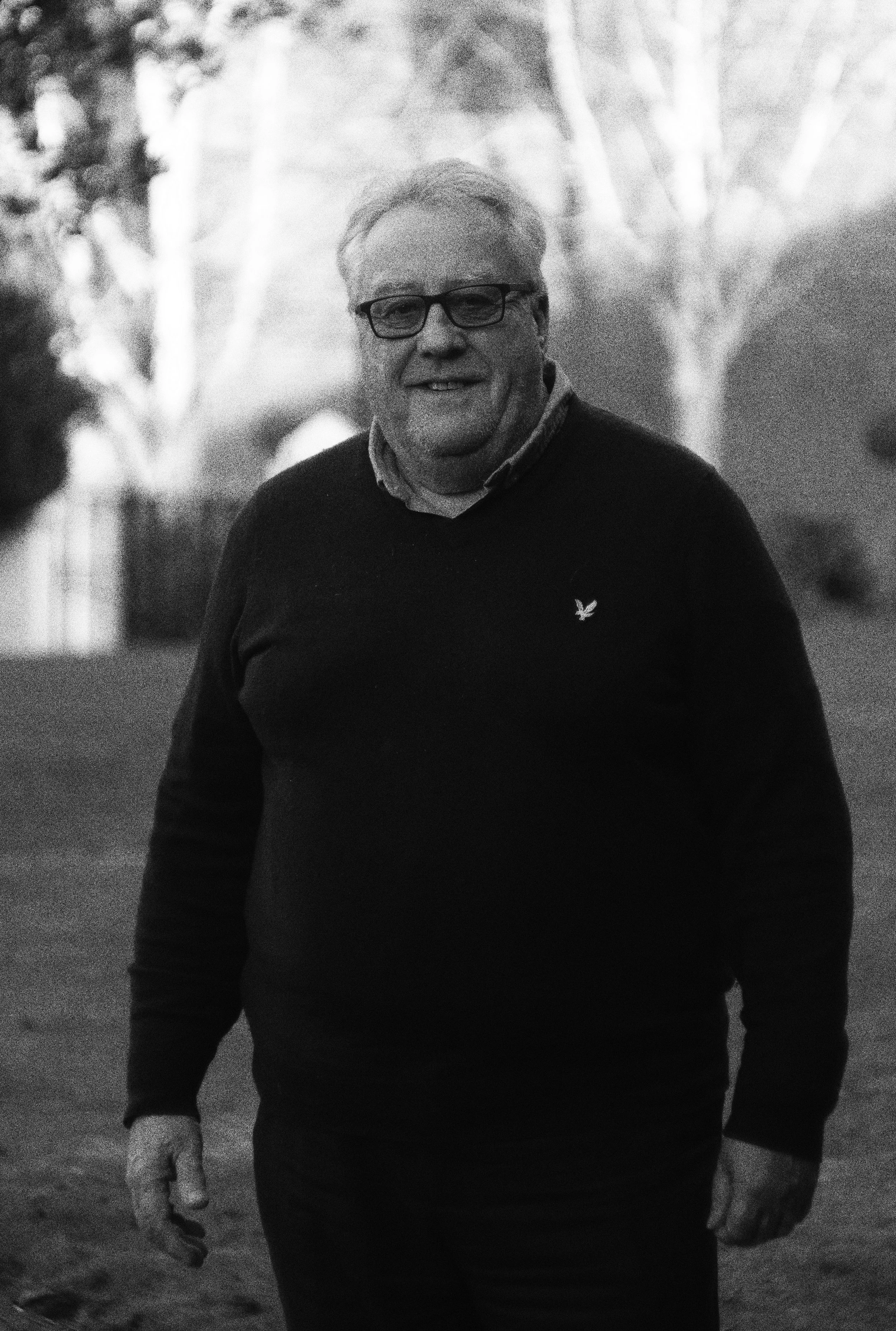 A smiling middle-aged man wearing glasses, a collared shirt, and a sweater with a small logo, standing outdoors in a park or yard with trees in the background.